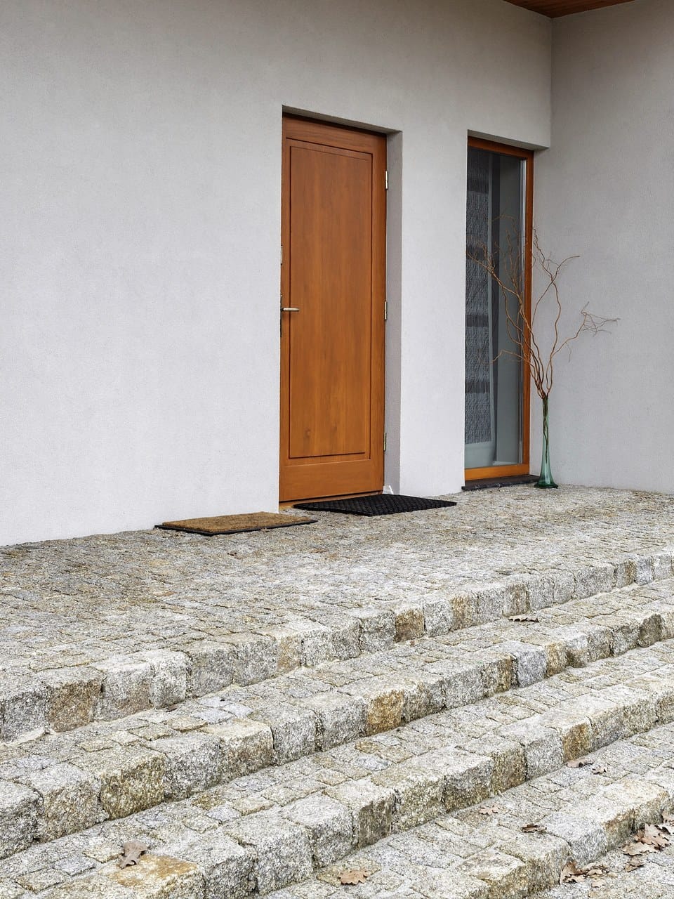 Porte d’entrée en bois naturel et haute fenêtre sur une maison contemporaine blanche, au-dessus de marches en granit